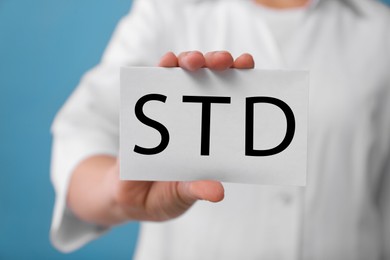 Photo of Doctor holding sheet of paper with abbreviation STD on light blue background, closeup