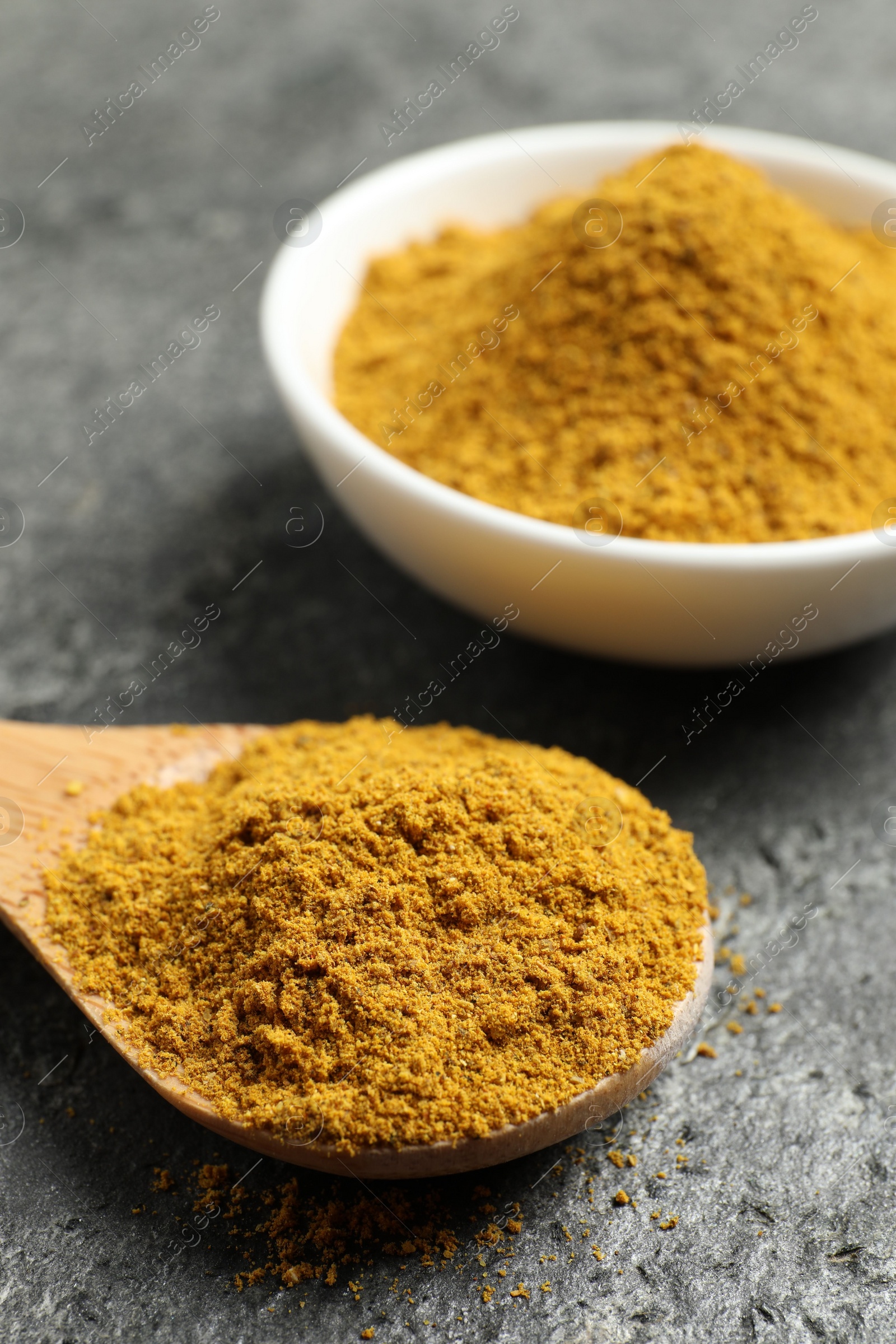 Photo of Spoon and bowl with dry curry powder on dark textured table, closeup