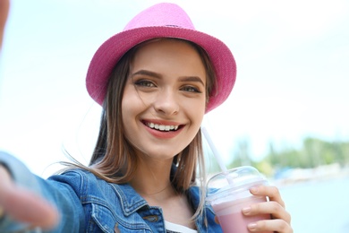 Photo of Happy young woman with drink taking selfie on riverside