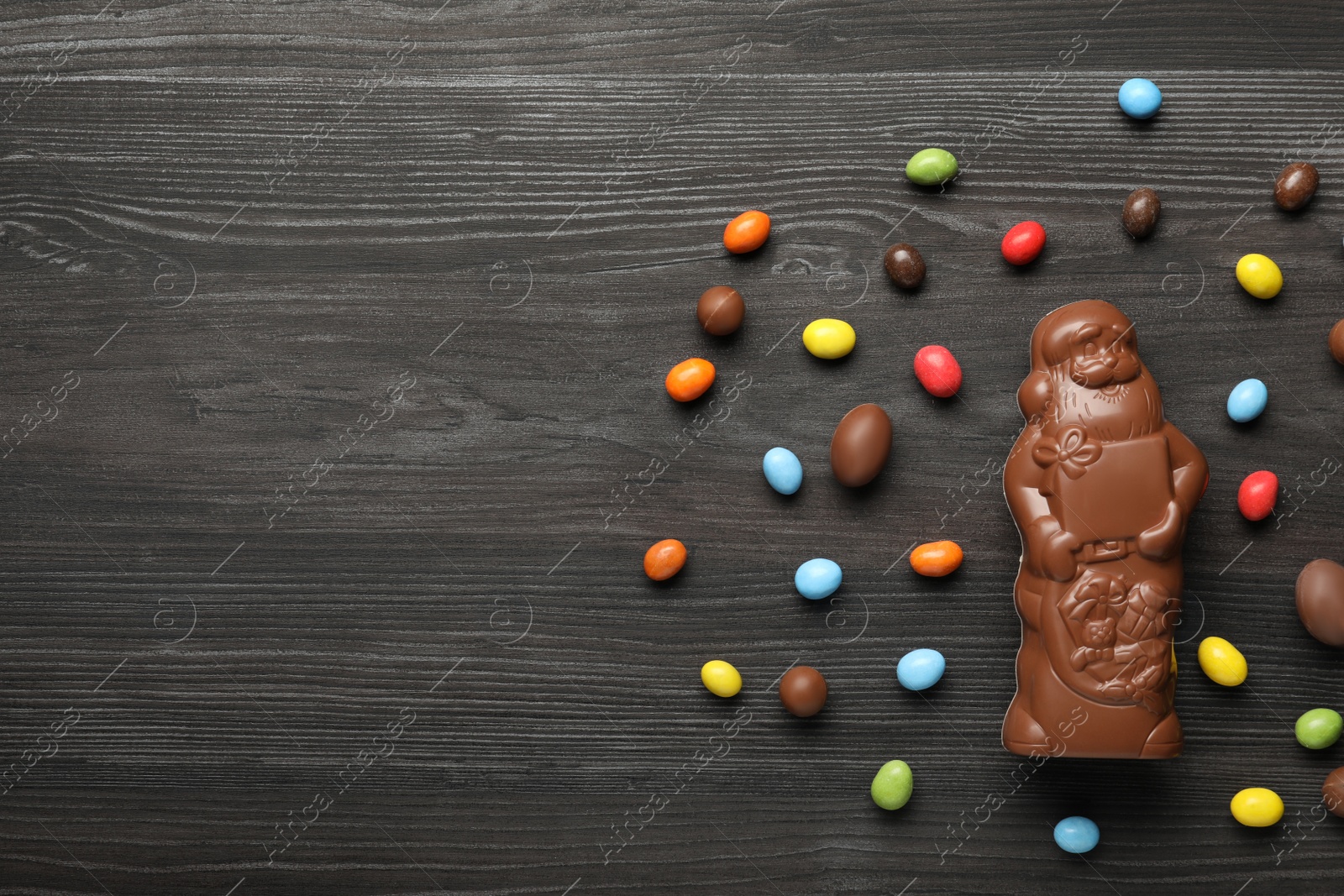 Photo of Flat lay composition of chocolate Santa Claus among sweets on dark grey wooden table, space for text