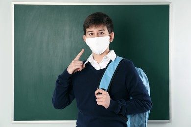 Photo of Boy wearing protective mask with backpack near chalkboard in classroom. Child safety