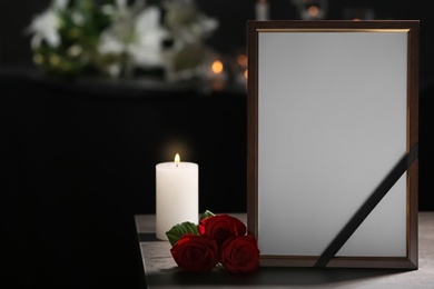 Funeral photo frame with black ribbon, roses and burning candle on table in dark room