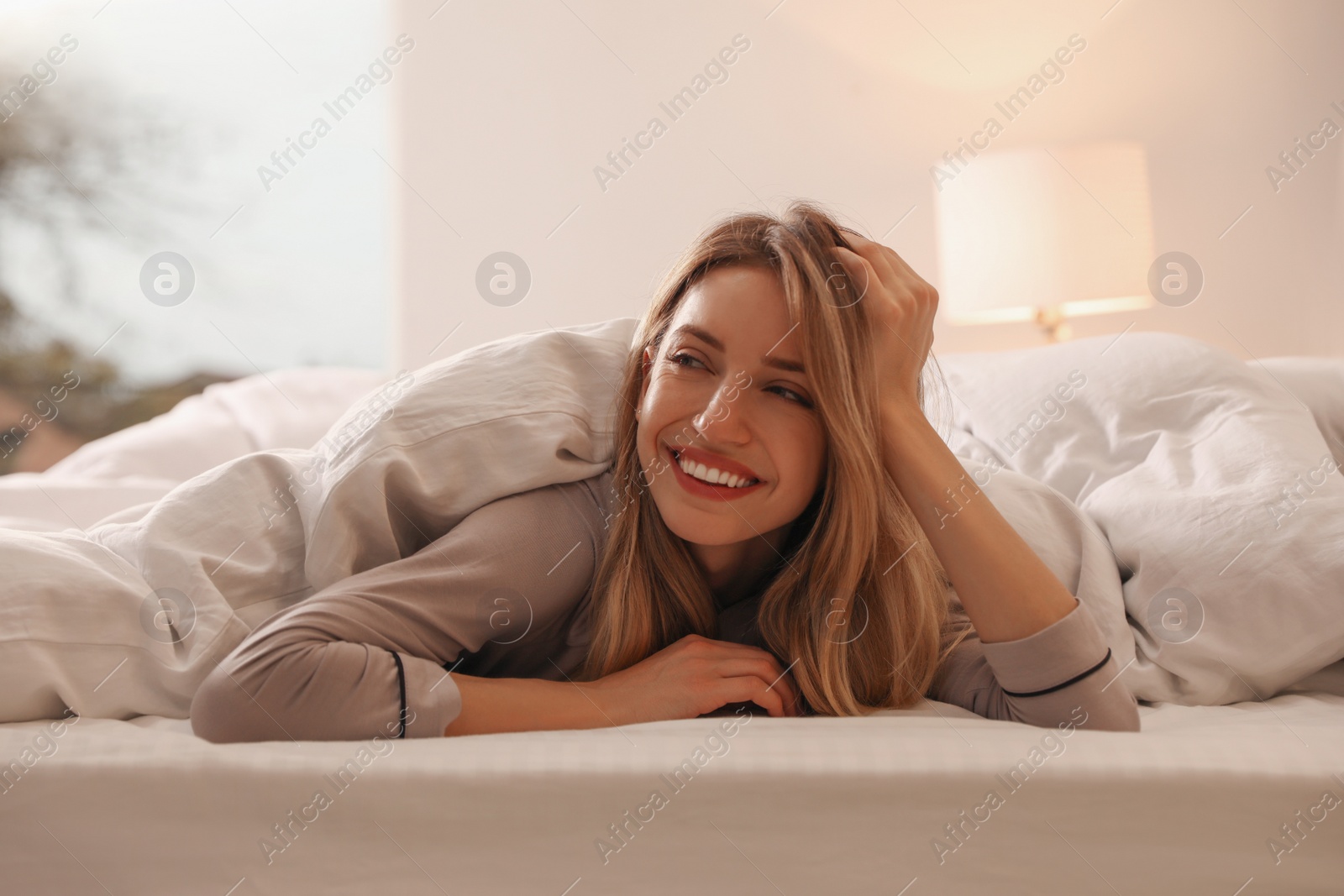 Photo of Cheerful woman covered with warm white blanket lying in bed indoors