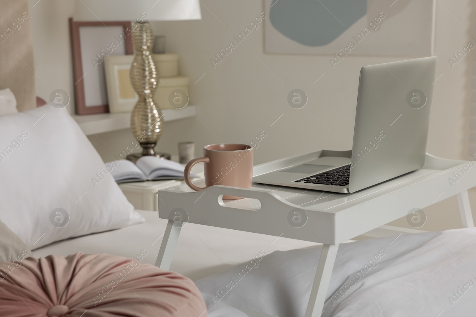 Photo of White tray table with laptop and cup of drink on bed indoors