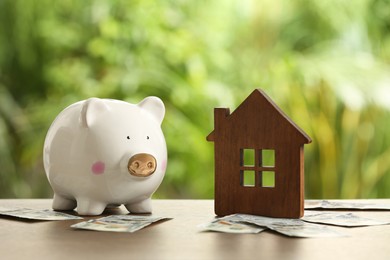 Photo of Piggy bank, house model and banknotes on wooden table outdoors