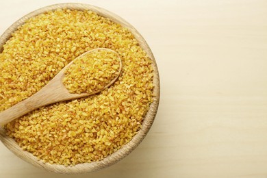 Bowl and spoon of uncooked bulgur on wooden table, top view. Space for text