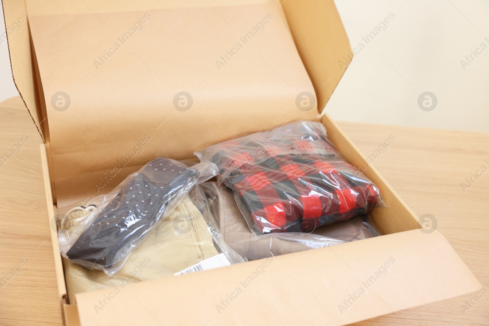 Photo of Open cardboard box with different clothes in plastic bags on wooden table. Delivery service