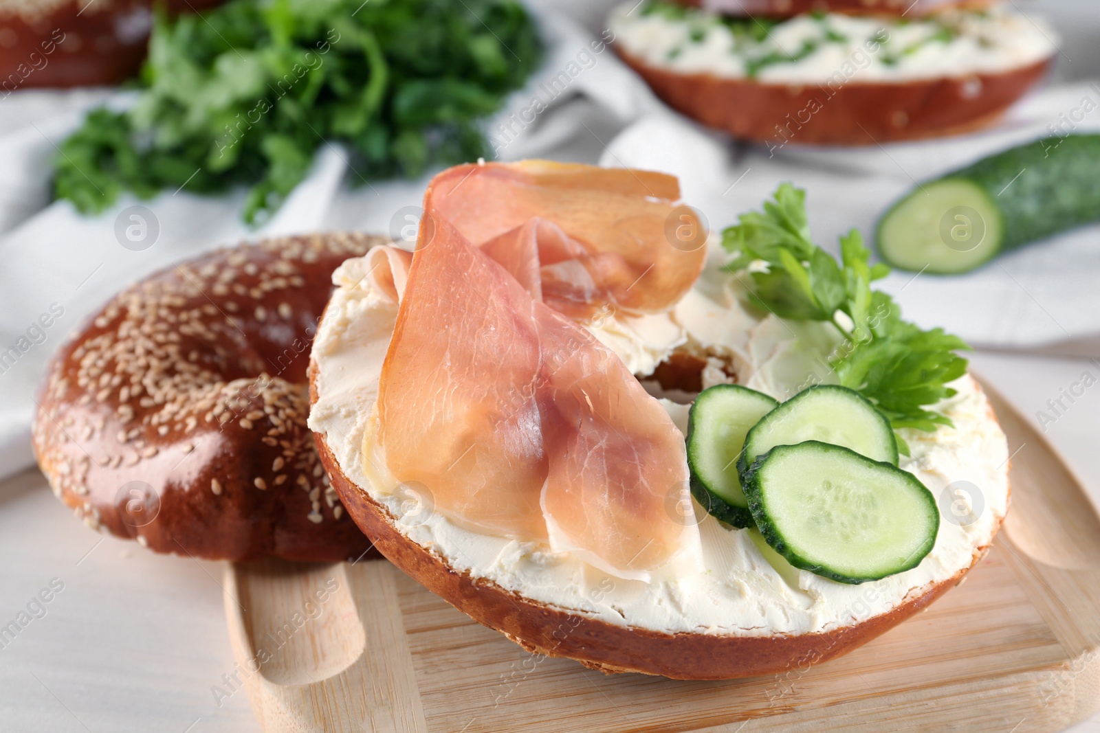 Photo of Delicious bagel with cream cheese, jamon, cucumber and parsley on wooden board, closeup