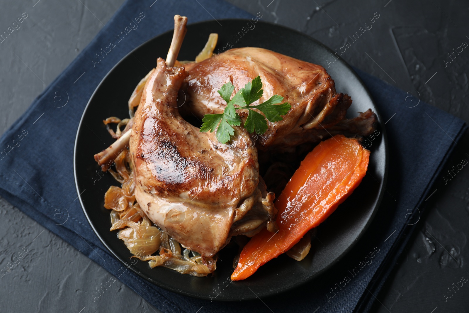 Photo of Tasty cooked rabbit meat with vegetables and parsley on black table, above view