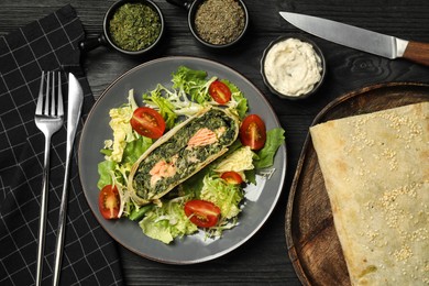Photo of Piece of tasty strudel with salmon, spinach and salad on black wooden table, flat lay
