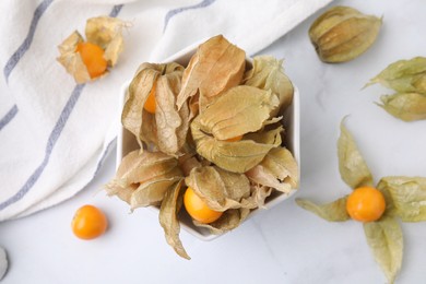 Photo of Ripe physalis fruits with calyxes in bowl on white marble table, flat lay