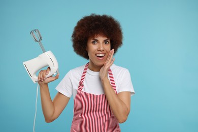 Photo of Emotional young woman in apron holding mixer on light blue background