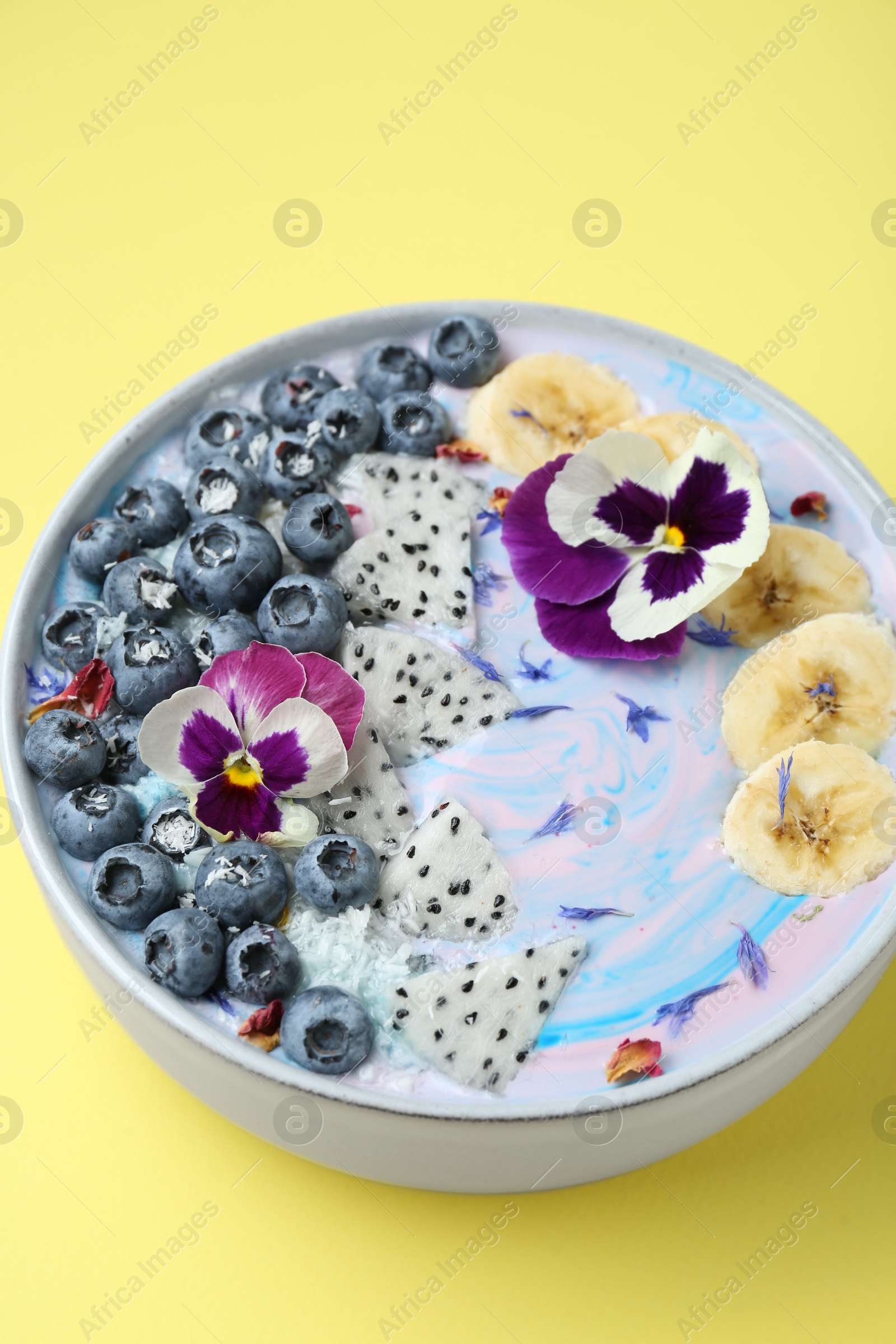 Photo of Delicious smoothie bowl with fresh fruits, blueberries and flowers on yellow background, closeup