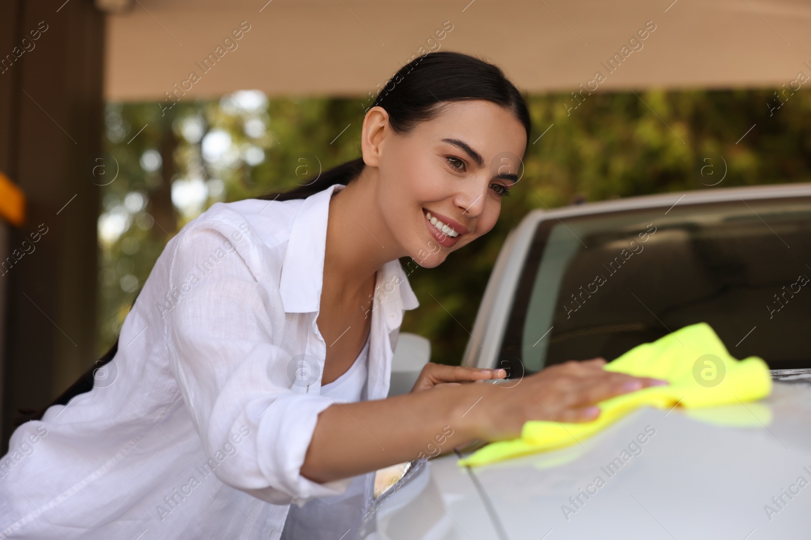 Photo of Happy woman cleaning car hood with rag outdoors