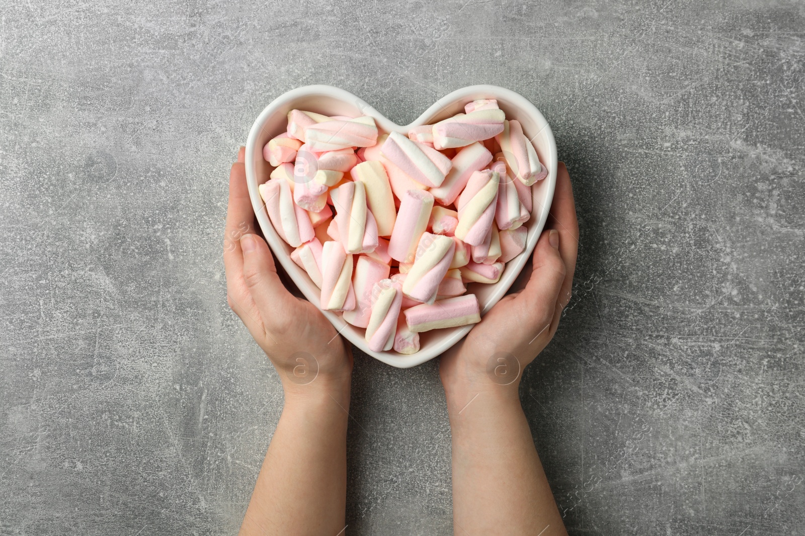 Photo of Woman holding heart shaped bowl full of marshmallows on grey background, top view