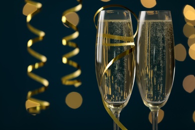 Photo of Glasses of champagne and serpentine streamer on dark blue background with blurred lights, closeup