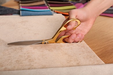 Photo of Man cutting beige fabric with scissors at wooden table, closeup