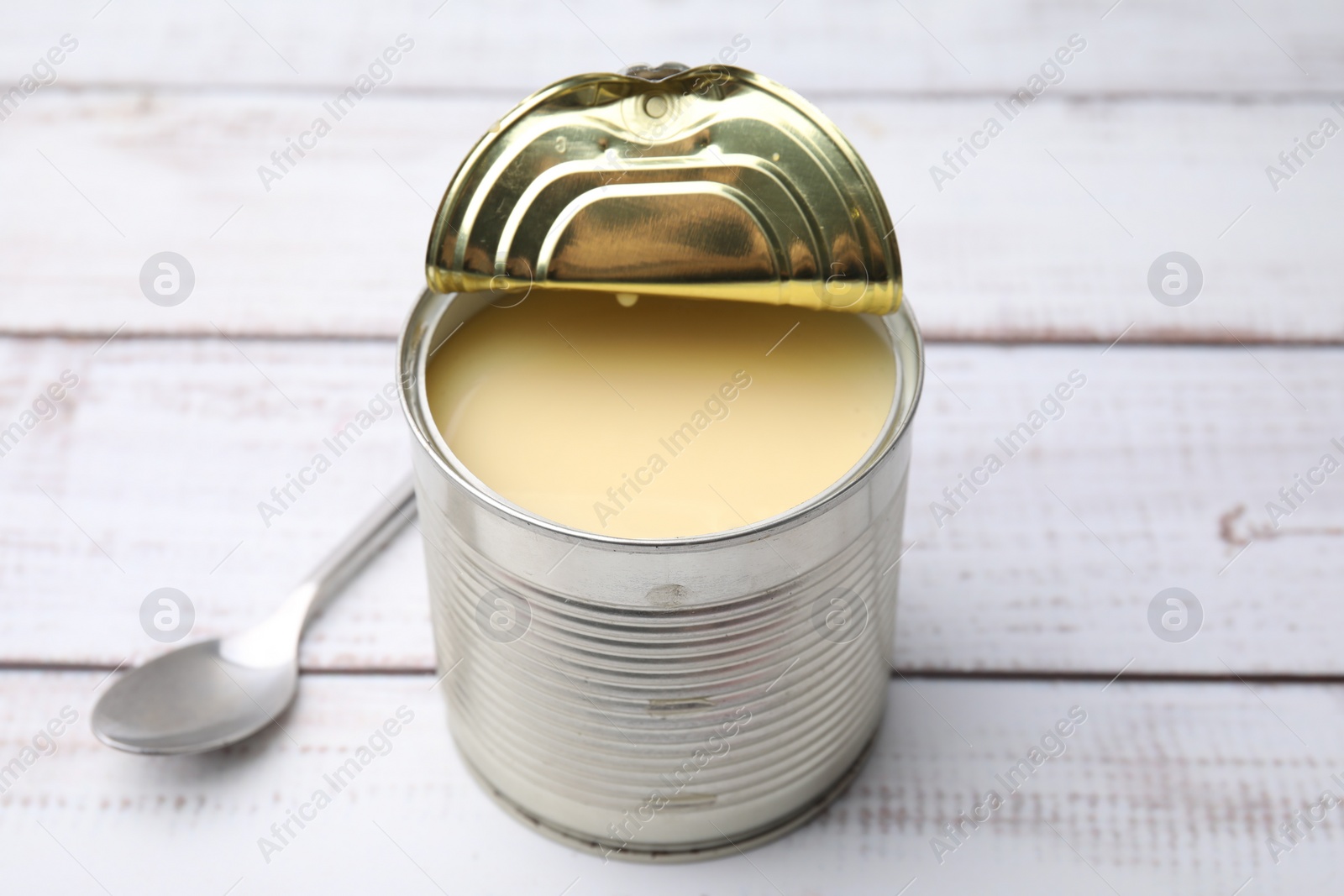 Photo of Tasty condensed milk in tin can and spoon on white wooden table, closeup