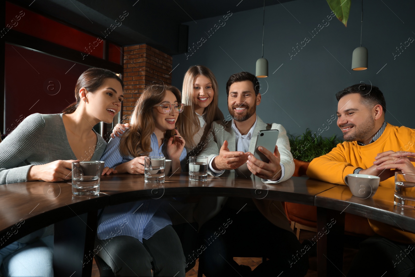 Photo of Handsome man showing something funny in smartphone to his friends in cafe