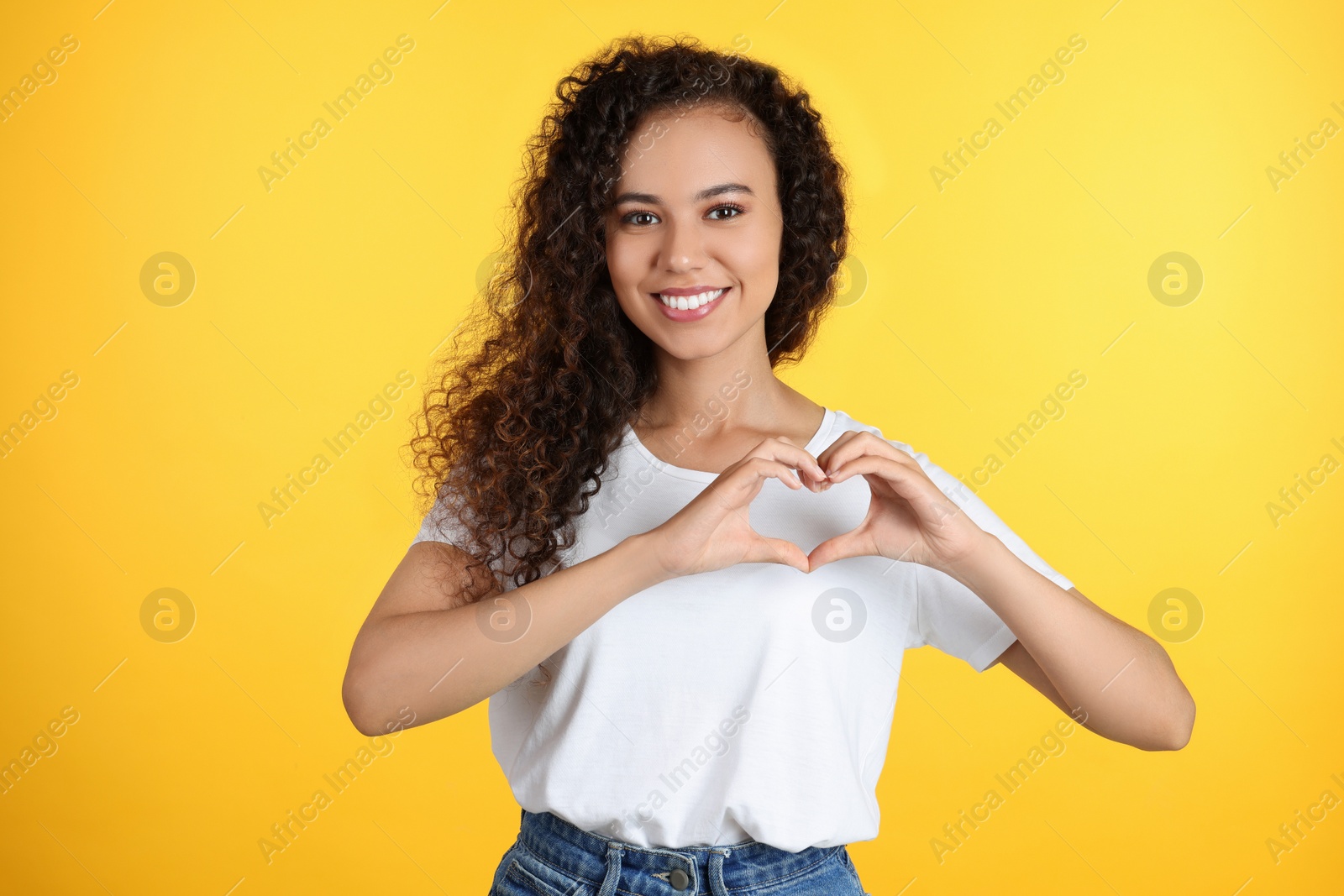 Photo of Happy young African-American woman making heart with hands on yellow background