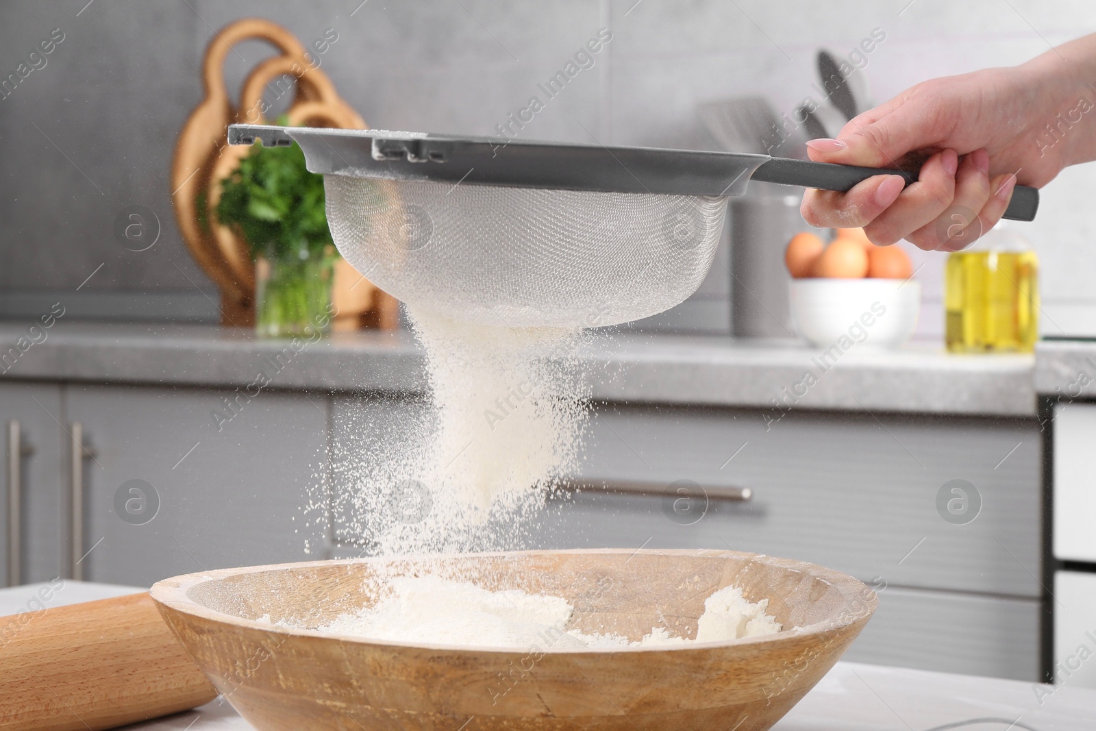 Photo of Woman sieving flour into bowl at white wooden table in kitchen, closeup