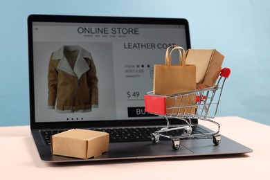 Photo of Online store. Laptop, mini shopping cart and purchases on beige table