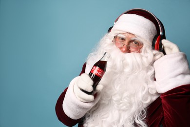 MYKOLAIV, UKRAINE - JANUARY 18, 2021: Santa Claus holding Coca-Cola bottle and listening to music with headphones on light blue background