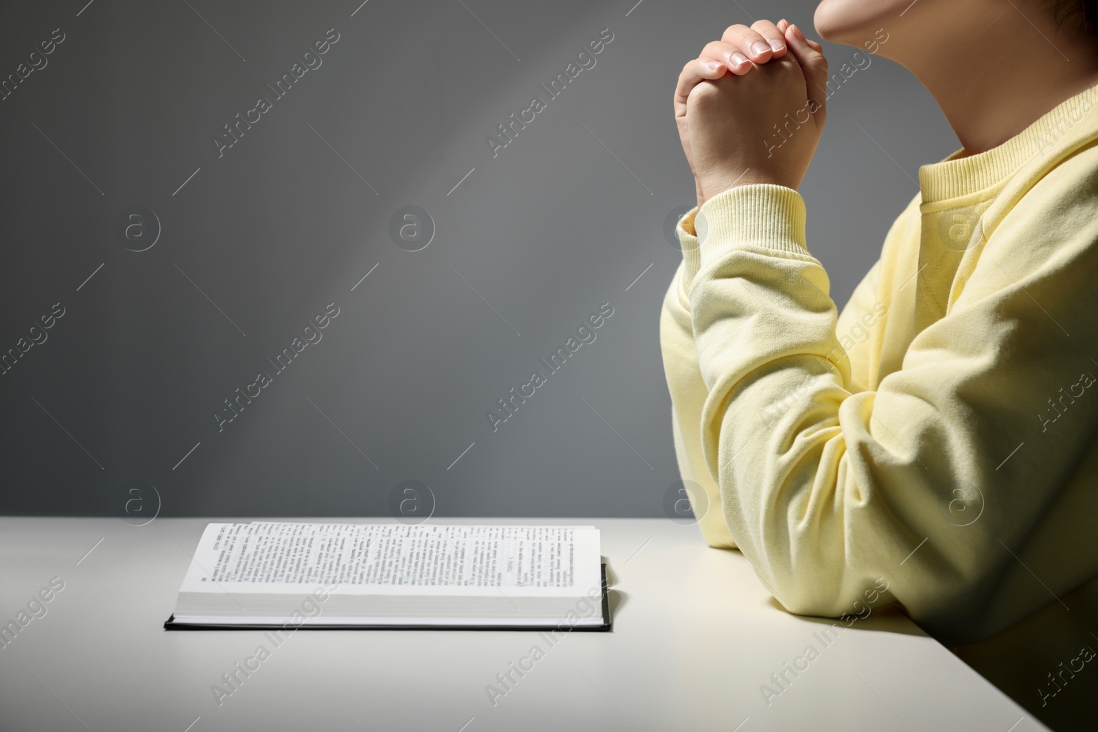 Photo of Woman praying over Bible at white table against grey background, closeup. Space for text