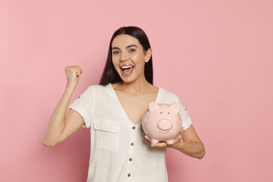 Photo of Emotional young woman with ceramic piggy bank on pale pink background