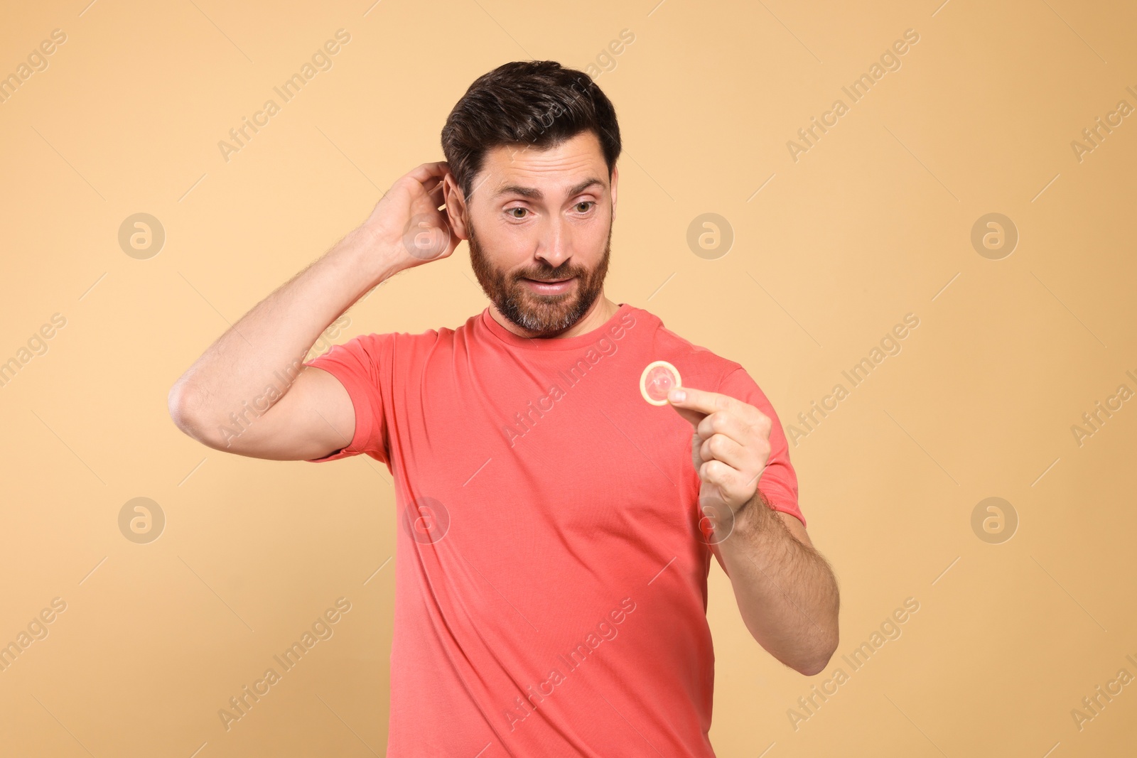 Photo of Confused man holding condom on beige background. Safe sex