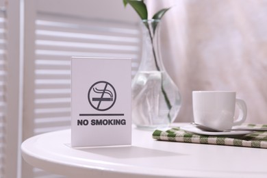 Photo of No Smoking sign and cup of coffee on white table indoors