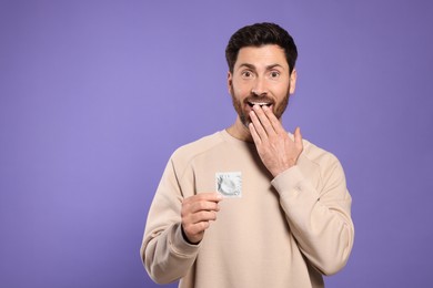 Emotional man holding condom on purple background, space for text. Safe sex