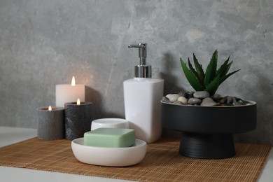 Photo of Potted artificial plant, soap, burning candles and bamboo mat on white table
