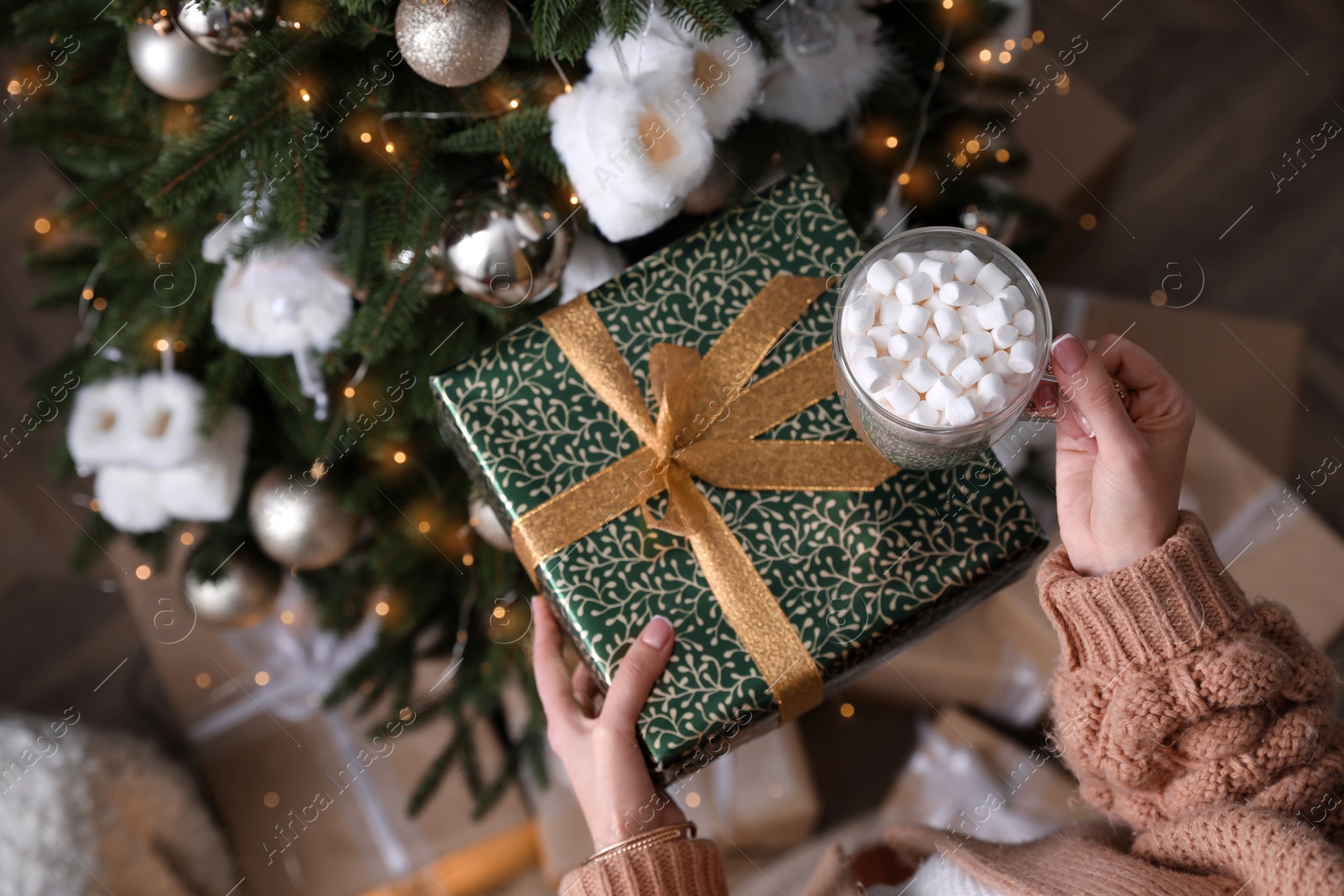 Photo of Woman with cup of delicious hot drink and gift box near Christmas tree at home, top view