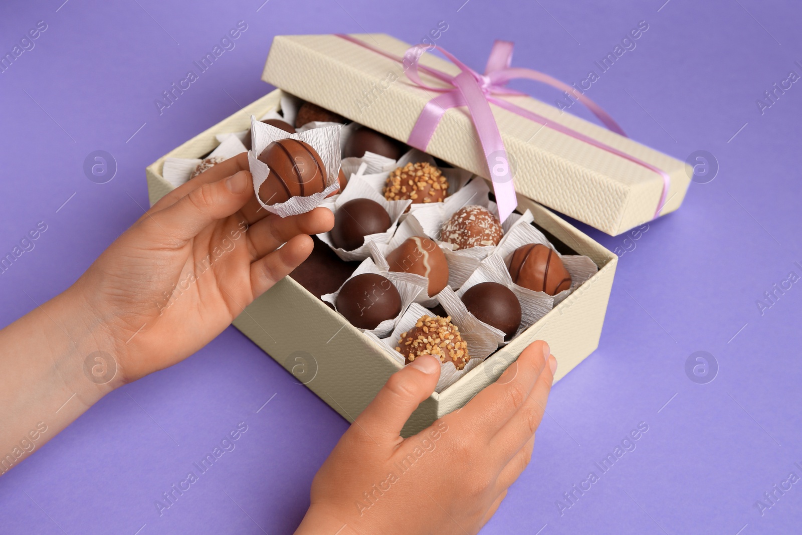 Photo of Child taking delicious chocolate candy from box on light purple background, closeup