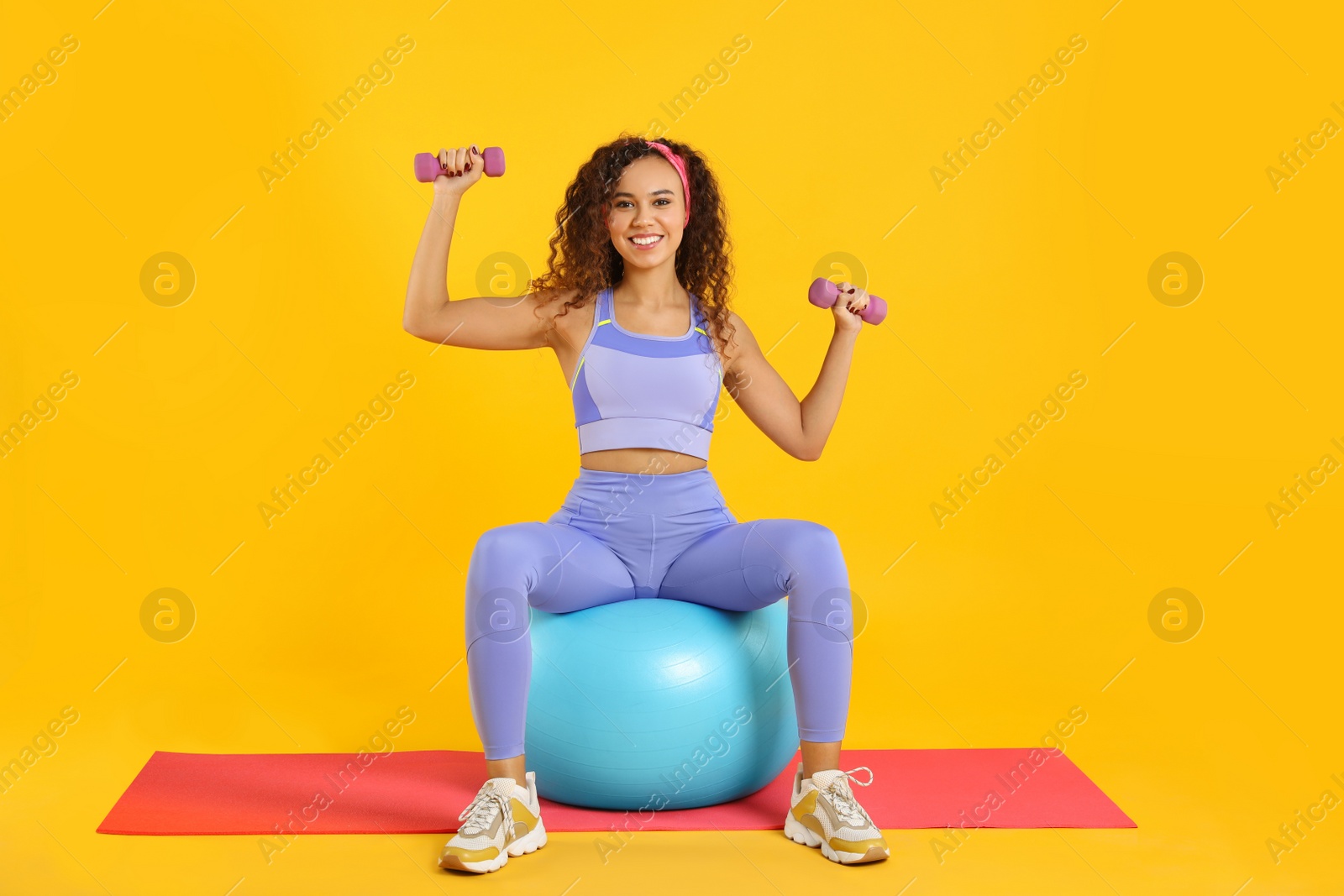 Photo of Beautiful African American woman doing exercise with fitness ball and dumbbells on yoga mat against yellow background
