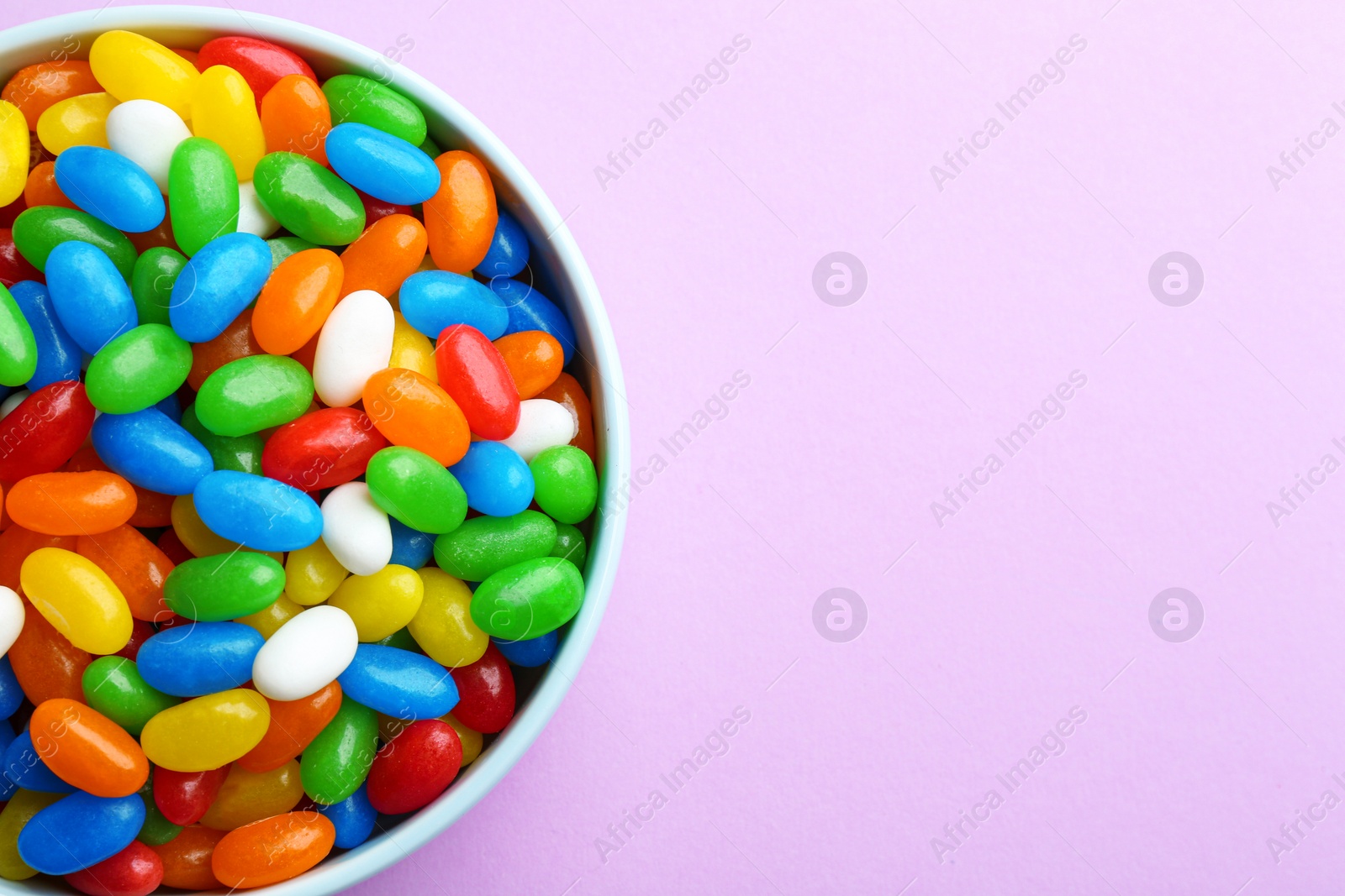 Photo of Bowl with colorful jelly beans on lilac background, top view. Space for text