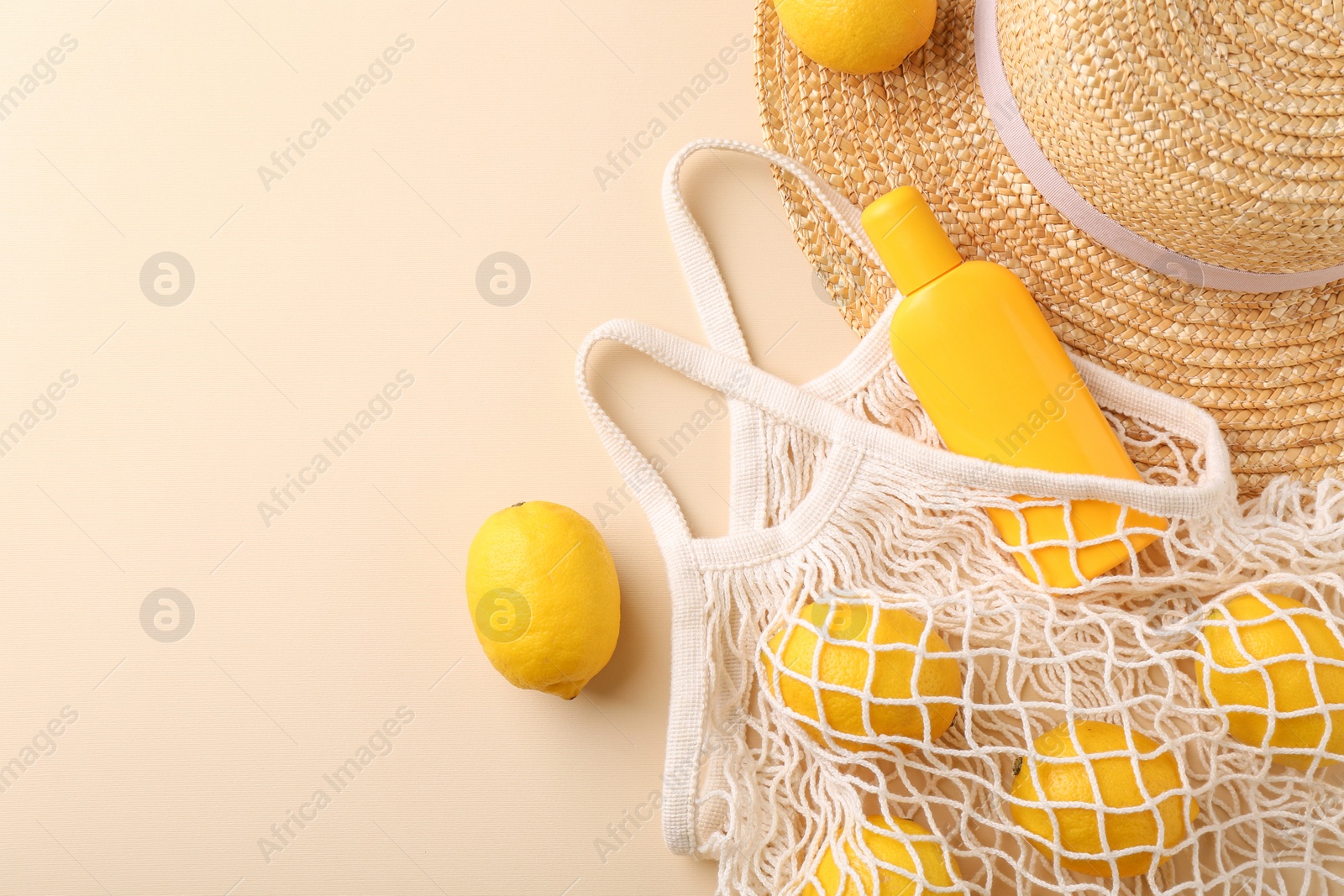 Photo of String bag with fresh lemons, sunscreen and straw hat on beige background, flat lay. Space for text