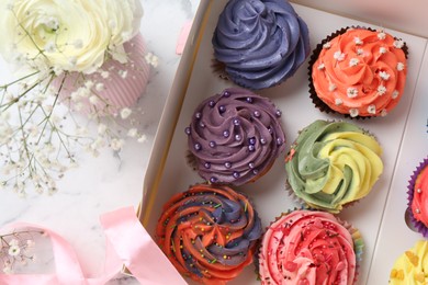 Different colorful cupcakes in box and flowers on white table, above view