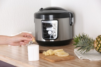 Photo of Woman putting pineapple into container with delicious fresh yogurt made in modern multi cooker on table, closeup