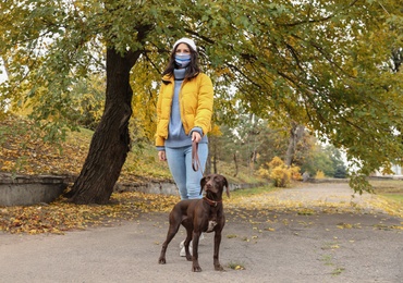 Photo of Woman in protective mask with German Shorthaired Pointer in park. Walking dog during COVID-19 pandemic