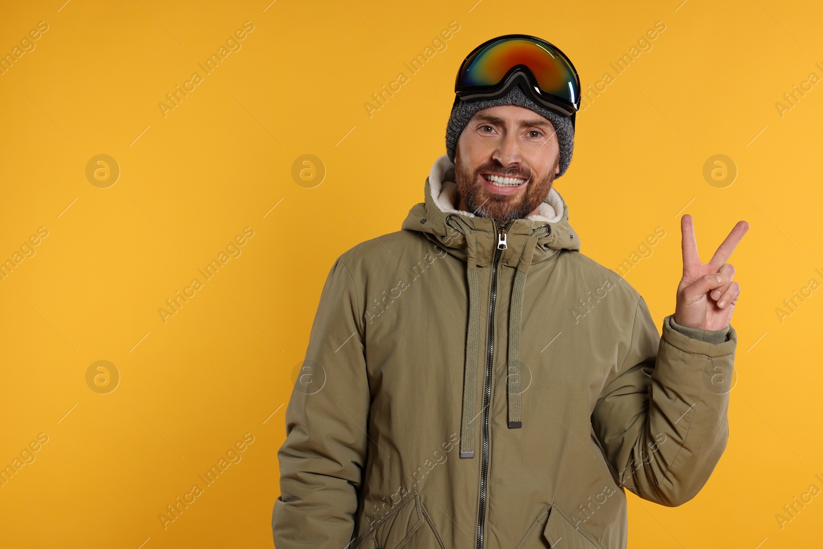 Photo of Winter sports. Happy man in ski suit and goggles showing V-sign on orange background, space for text