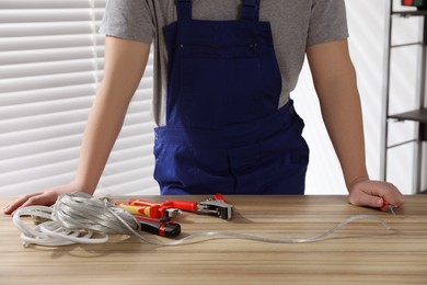 Photo of Professional electrician in uniform, tools and wire at wooden table indoors, closeup