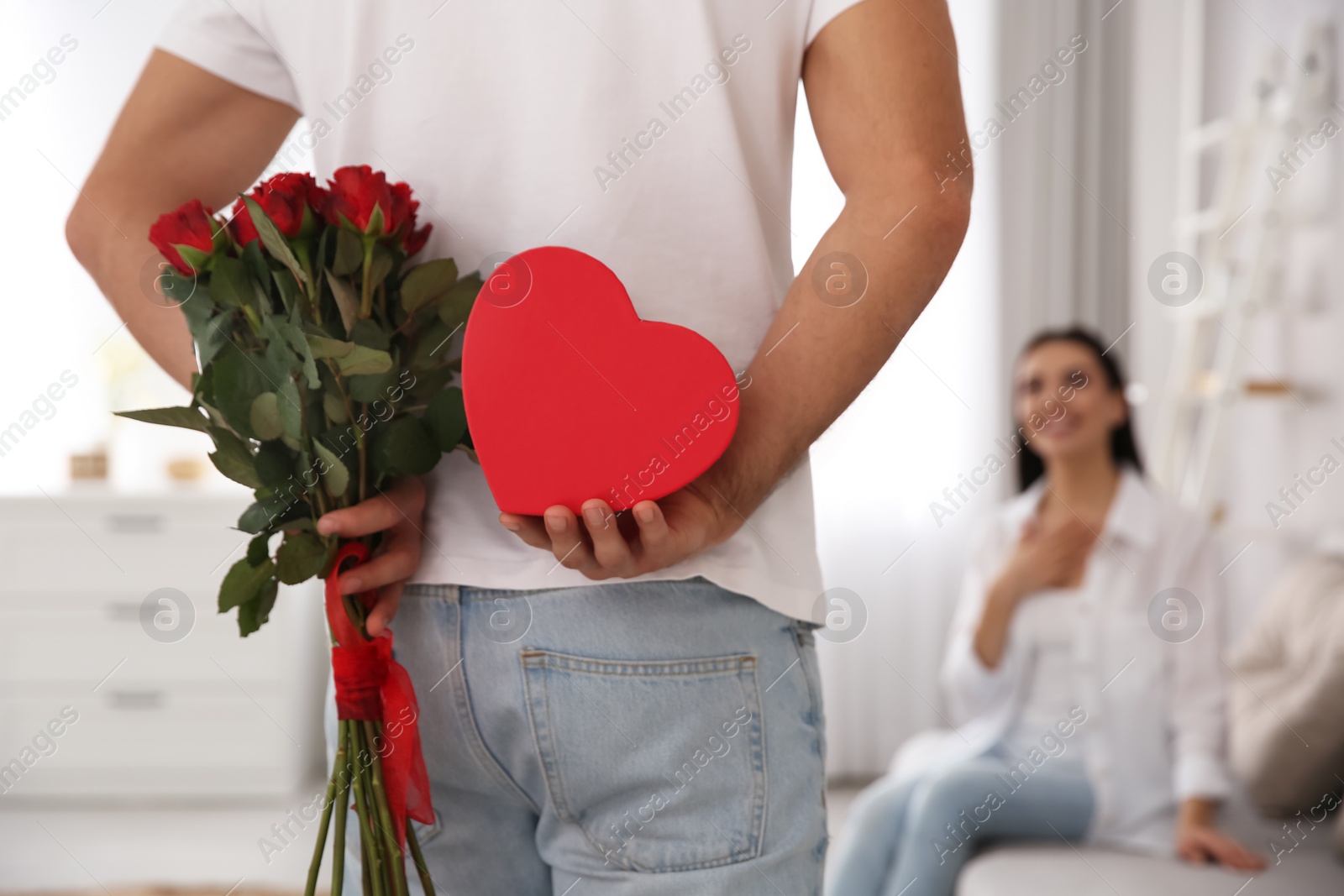 Photo of Man hiding beautiful bouquet and gift box behind his back at home, closeup. Valentine's day celebration