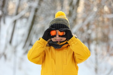 Photo of Cute little boy covering eyes with tangerines in snowy park on winter day