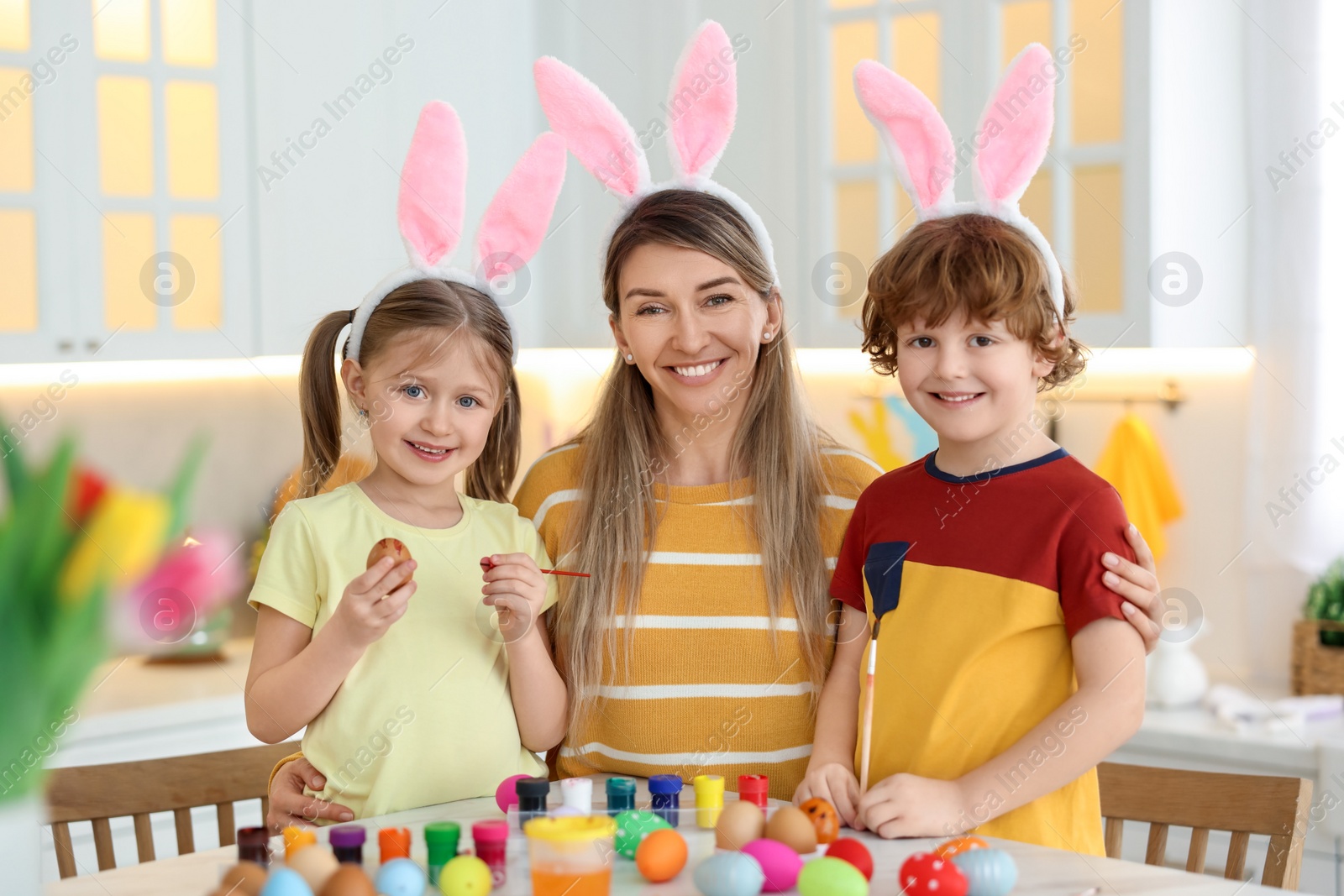 Photo of Painting Easter eggs. Happy mother and her cute children with bunny ears at table in kitchen