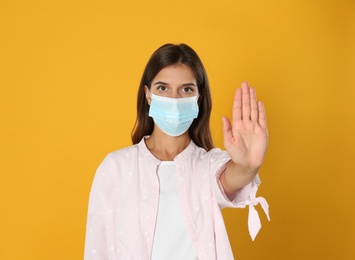 Photo of Woman in protective mask showing stop gesture on yellow background. Prevent spreading of coronavirus