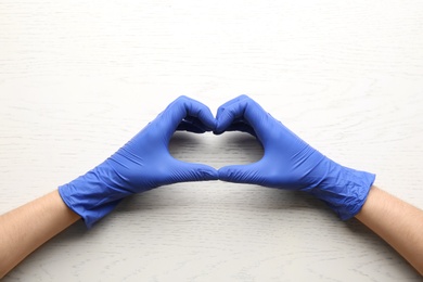 Person in medical gloves showing heart gesture on white wooden background, closeup of hands