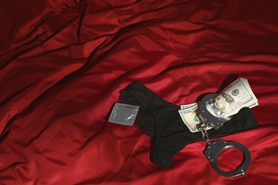 Photo of Prostitution concept. Black panties, dollar banknotes, condom and handcuffs on red fabric, space for text