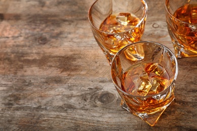 Golden whiskey in glasses with ice cubes on wooden table. Space for text